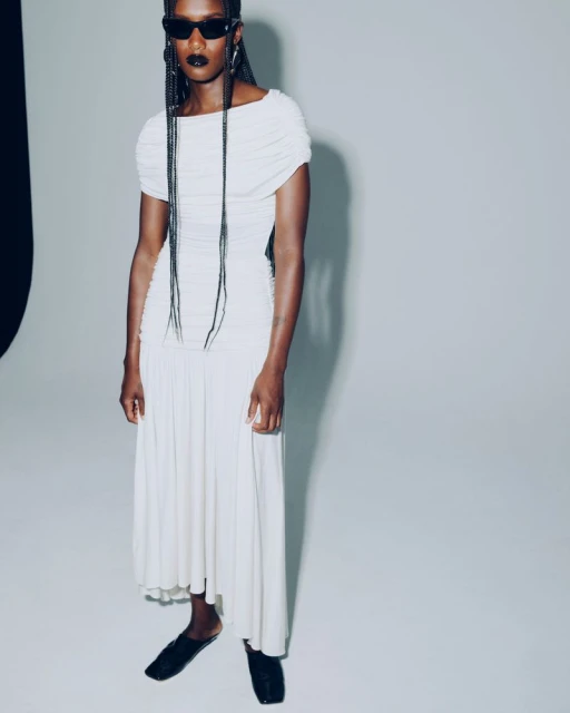 An African American woman models a white dress and black flats in a studio. She wears black sunglasses, and her braids hang long.