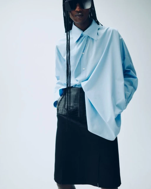 Fashion shot of a Black model in a light blue shirt and black skirt, posing in a photo studio against a white backdrop.