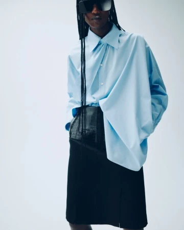 Fashion shot of a Black model in a light blue shirt and black skirt, posing in a photo studio against a white backdrop.