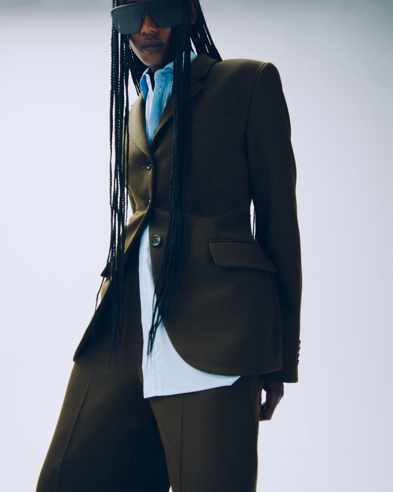 Woman in tailored olive-green suit with wide-leg trousers and visor sunglasses against white backdrop, studio lighting, fashion editorial.