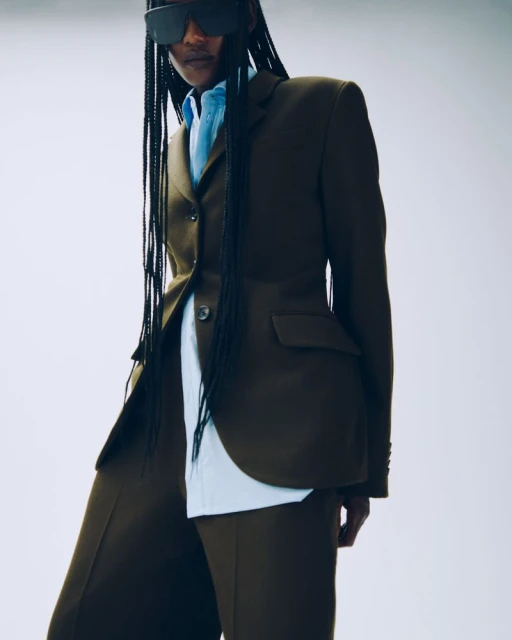 Woman in tailored olive-green suit with wide-leg trousers and visor sunglasses against white backdrop, studio lighting, fashion editorial.