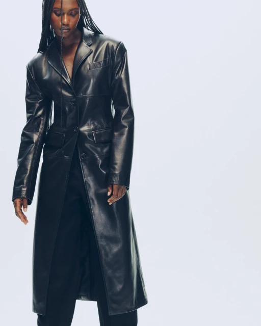 A slender Black woman in a black leather coat poses in a studio. Fashion photography with a bright white background and professional lighting.