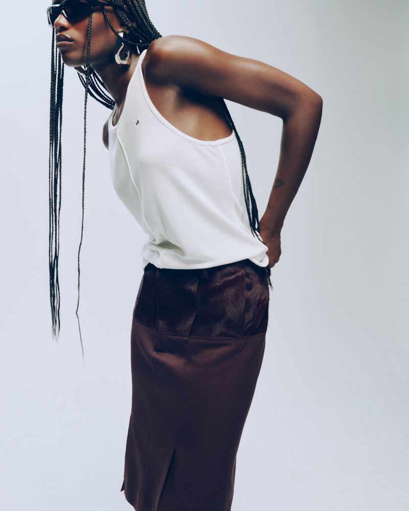 Model Essence Taylor in a white tank top, dark brown skirt, and sunglasses posing against a seamless studio backdrop.