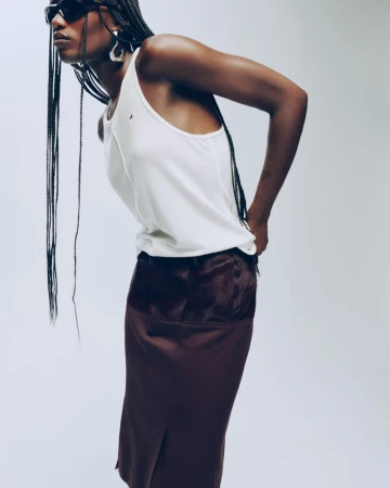 Model Essence Taylor in a white tank top, dark brown skirt, and sunglasses posing against a seamless studio backdrop.