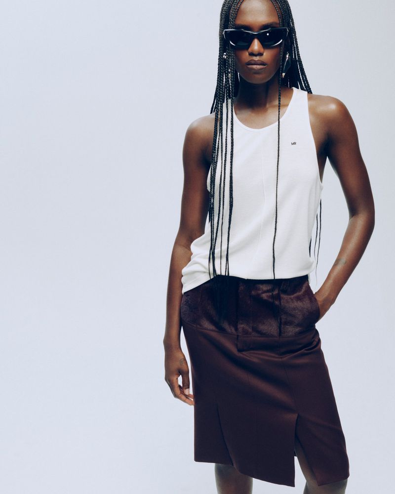 Essence Taylor wearing a white tank top, dark burgundy skirt, and black sunglasses against a white studio background.