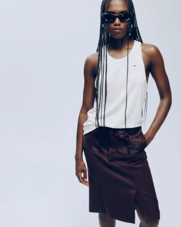 Essence Taylor wearing a white tank top, dark burgundy skirt, and black sunglasses against a white studio background.