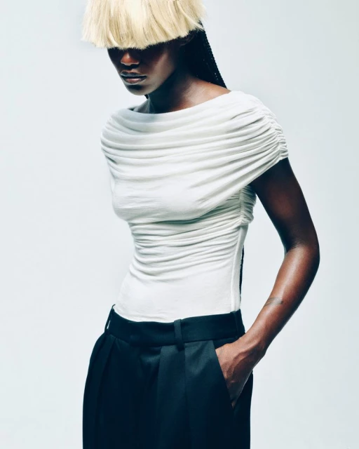 Fashion portrait of a Black woman in a white draped top and black trousers against a bright background, with unique blonde fringe.