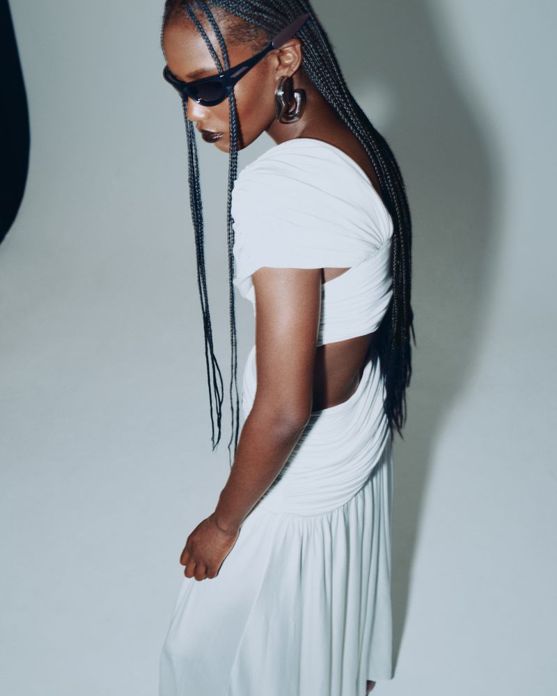 Essence Taylor wearing a white draped cut-out dress, dark sunglasses, and long braids against a bright studio background.