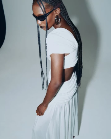 Essence Taylor wearing a white draped cut-out dress, dark sunglasses, and long braids against a bright studio background.