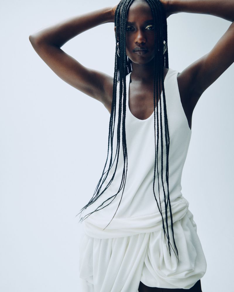 Portrait of an African woman with long braids in a studio setting. She poses with her hands behind her head, wearing a white outfit.