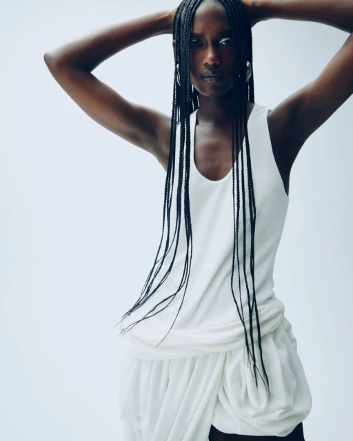 Portrait of an African woman with long braids in a studio setting. She poses with her hands behind her head, wearing a white outfit.