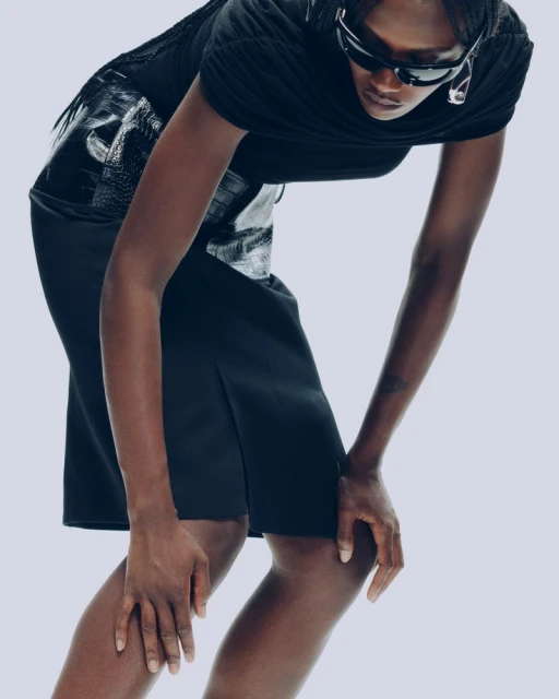 A black woman models black skirt and top, leaning forward, shot in photo studio with gray backdrop and dramatic lighting in vertical fashion shot.