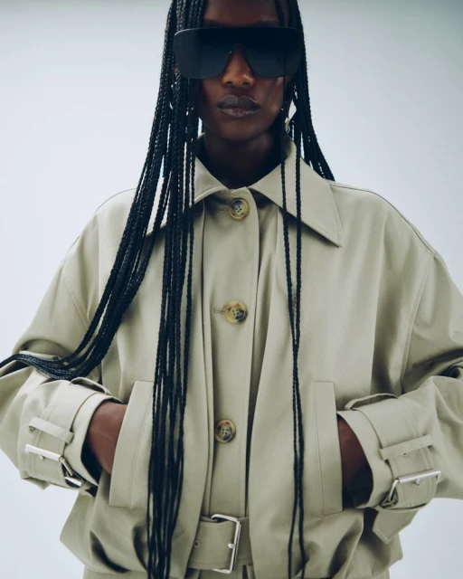 An adult female model with braided hair wearing large sunglasses and a tan-colored trench coat is featured in a studio shot against a white background.