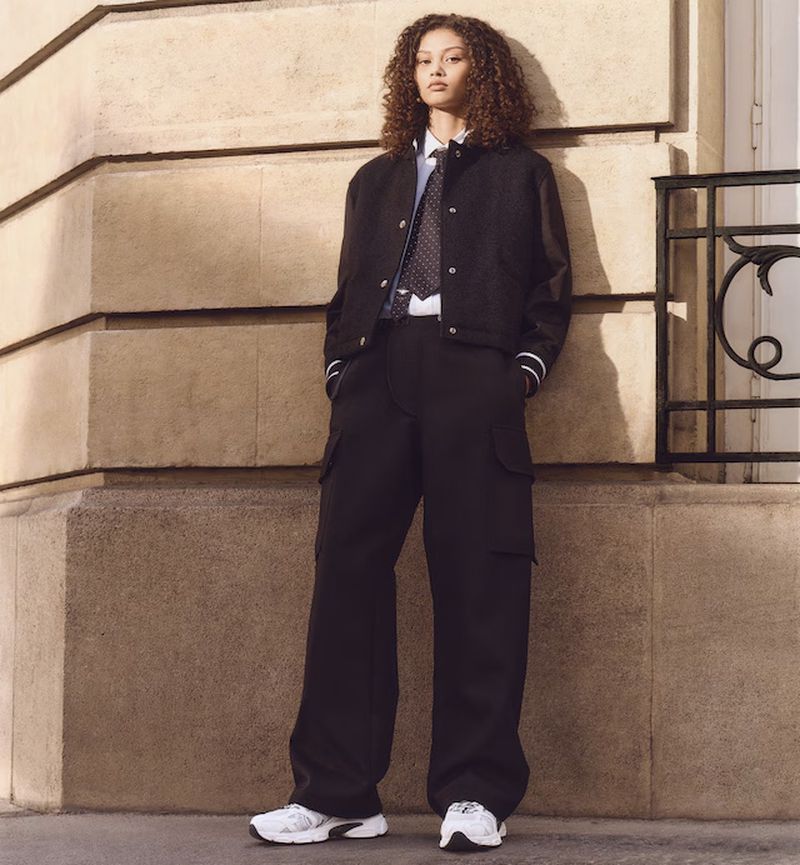 Fashionable woman in a black bomber jacket and wide-leg cargo pants, posing against a textured wall with a decorative iron railing.