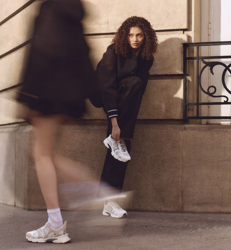 Woman in black suit ties white sneakers. Second blurred woman walks by on city street with beige wall in outdoor fashion shot.