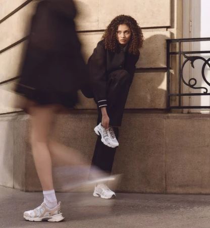 Woman in black suit ties white sneakers. Second blurred woman walks by on city street with beige wall in outdoor fashion shot.