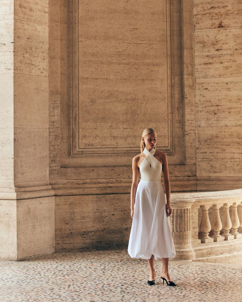A full shot of a blonde adult female fashion model in a white dress with black shoes, posing in a beige colonnade setting.