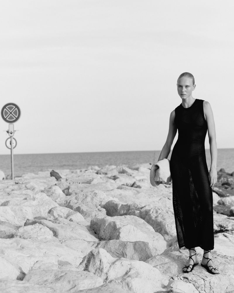 A woman in a black dress stands on a rocky shore near the sea, with a stop sign visible in the background. Black and white photography.