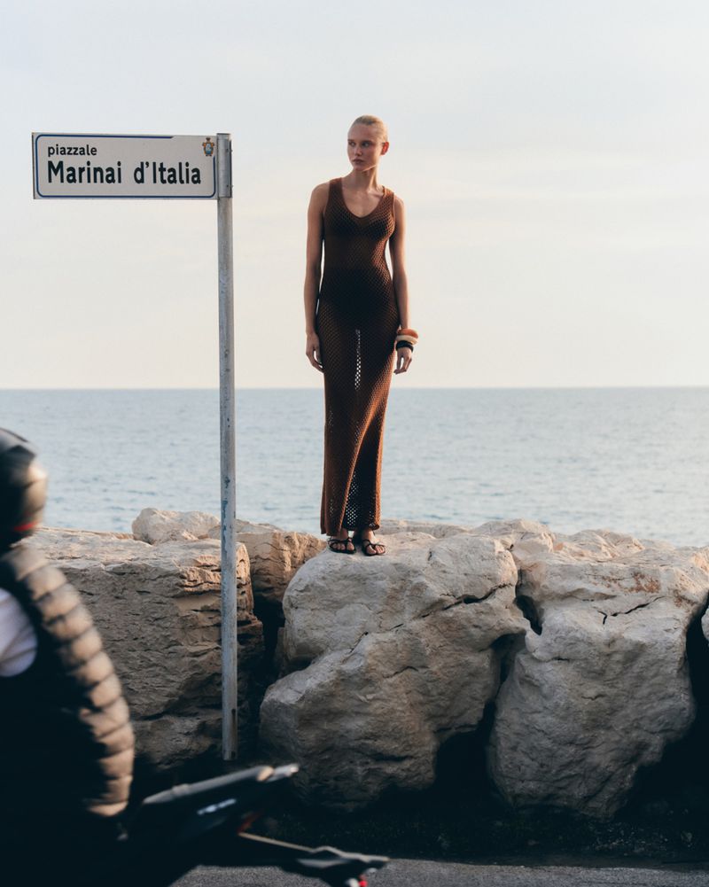 A slender fashion model in a brown maxi dress strikes a pose on a stone ledge near the sea, with a blurred motorcyclist in the foreground.