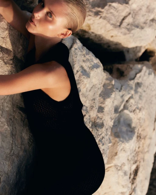 Blonde woman wearing a black knit dress leans against a rock formation; soft, warm light on her face and body in an outdoor setting.