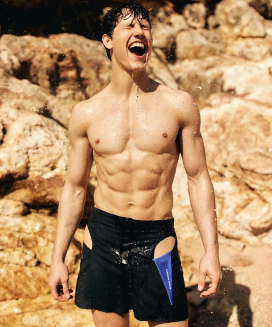 Muscular man laughing on a beach in black shorts with cutouts, wet from water with tan rock formations in the background.