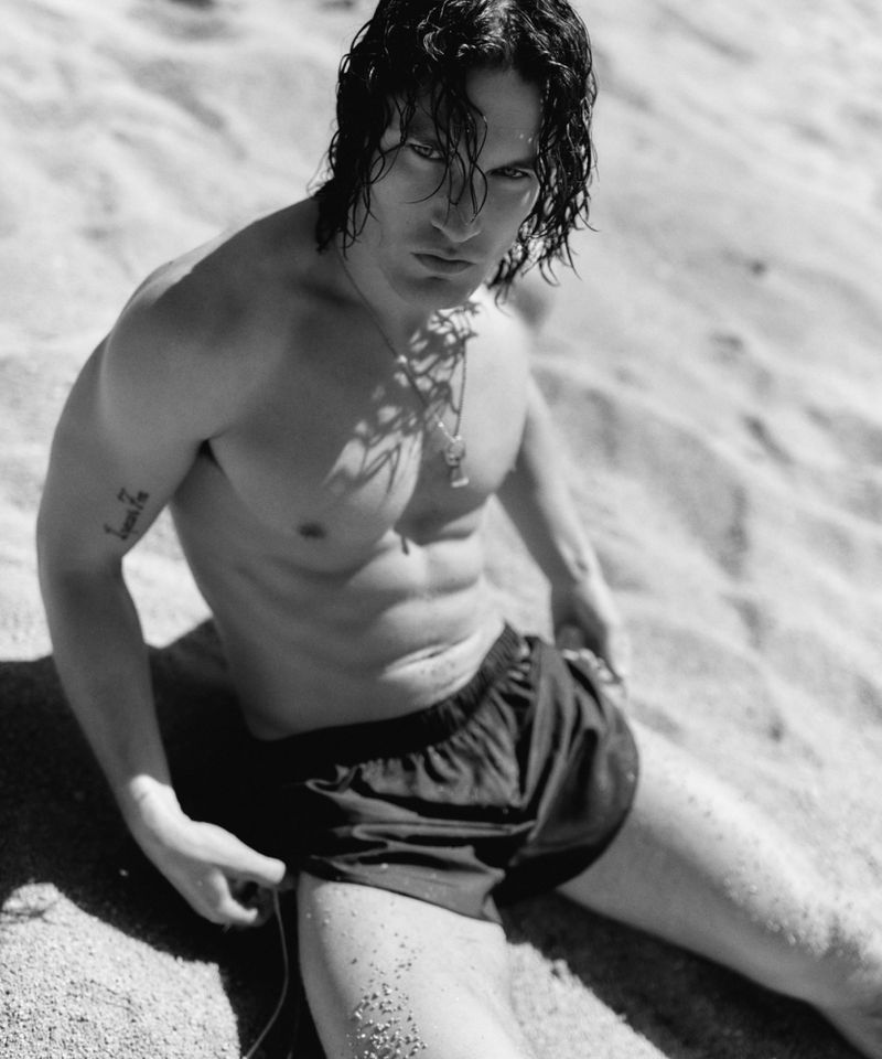 Monochrome portrait of muscular male model with dark hair posing shirtless on a sandy beach in black shorts looking at the camera.