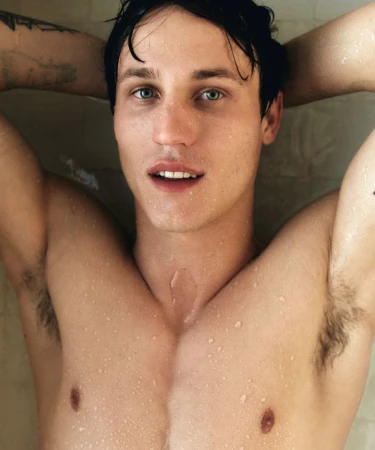 Close-up portrait of a shirtless young man with wet skin and hair resting his arms behind his head against a tiled wall.