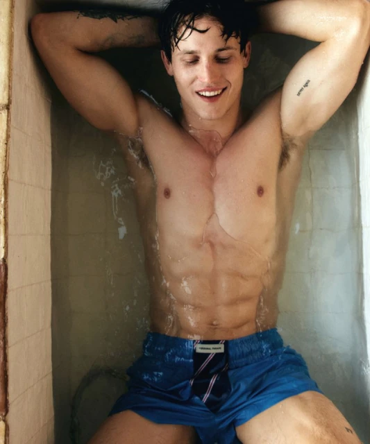 Smiling adult male model in a bathtub with water wearing blue swim trunks with tie detail, indoors with neutral wall tiles.