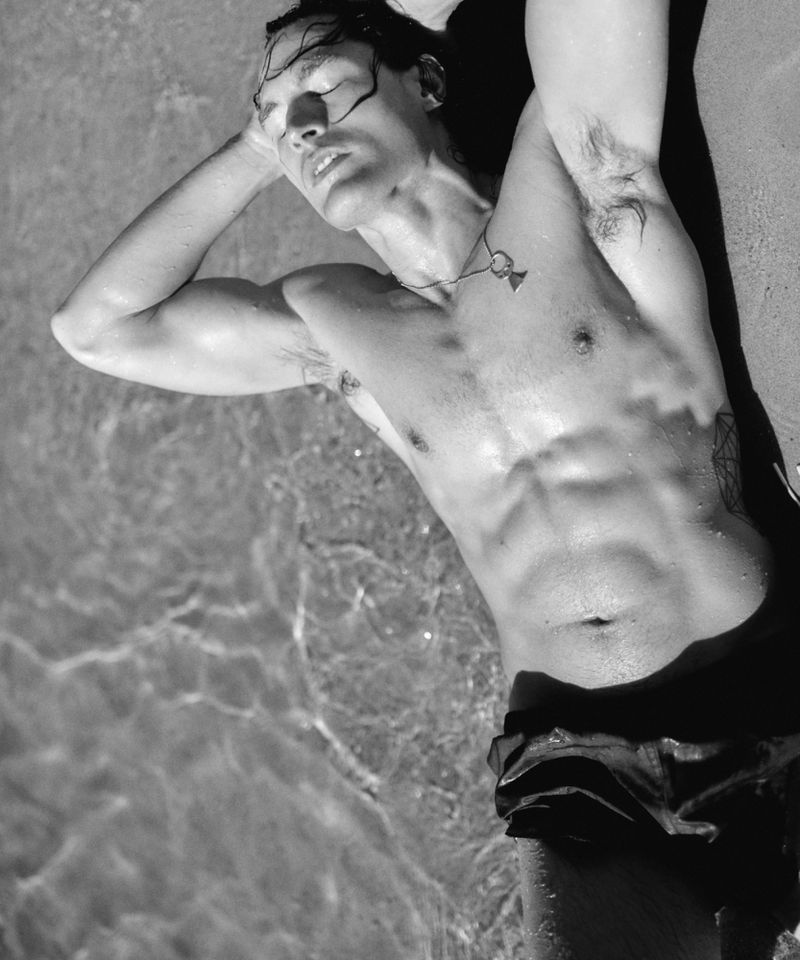 Black and white photo of a shirtless man lying back in shallow water on a sandy beach with hands behind his head.