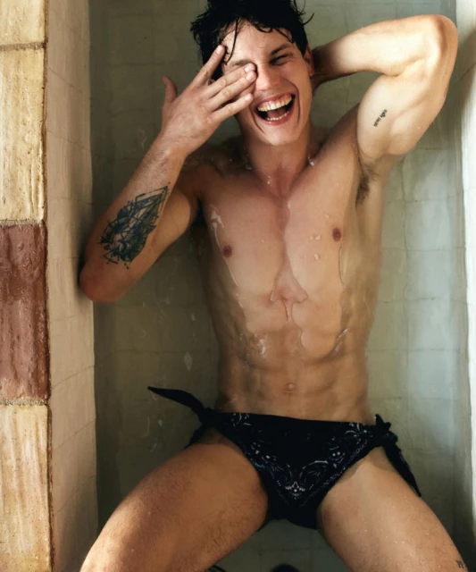 A shirtless man with tattoos smiles in the shower, covering one eye with his hand, wearing a black patterned speedo with a tie.