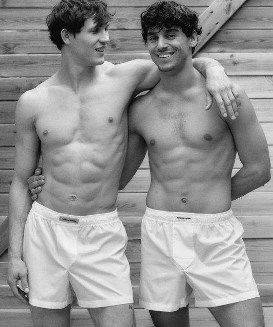 Two athletic adult men pose shirtless in white boxer shorts against a wood panel background, showcasing fitness and male fashion.
