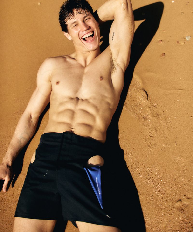 Fit adult male poses shirtless on a sandy beach, showing abs, wearing black shorts with side cut-outs, and smiling broadly.