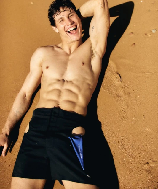 Fit adult male poses shirtless on a sandy beach, showing abs, wearing black shorts with side cut-outs, and smiling broadly.