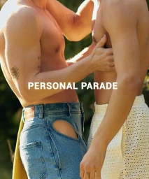 Two shirtless men standing closely together outdoors, one wearing cut-out blue jeans and the other in patterned white shorts.