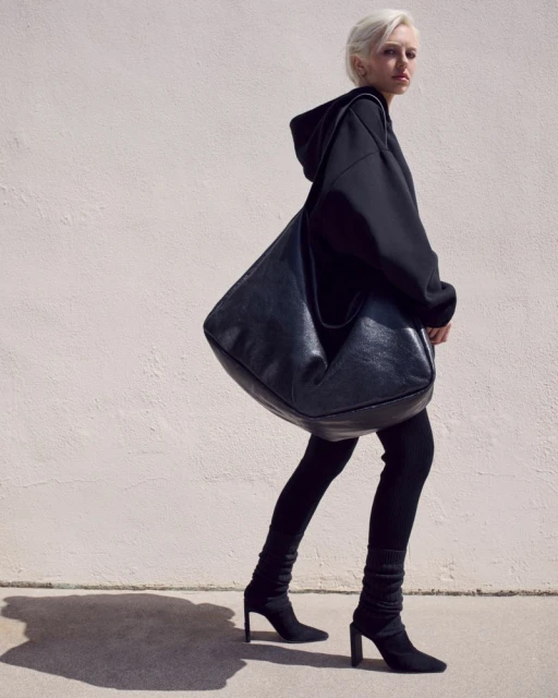 A woman walks toward the camera wearing all black with a large leather bag over her shoulder in a brightly lit outdoor setting.