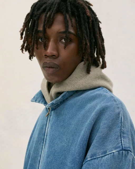 Close-up portrait of a young African-American man with dreadlocks wearing a denim jacket over a beige hoodie indoors.