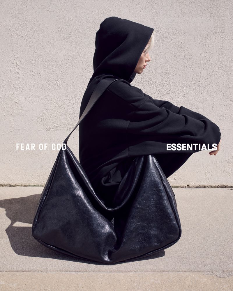 Woman in a black hooded sweatshirt sitting next to a large black leather hobo bag against a neutral wall backdrop.