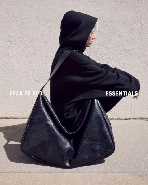 Woman in a black hooded sweatshirt sitting next to a large black leather hobo bag against a neutral wall backdrop.