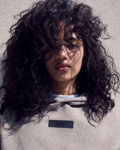 Close-up shot of a young adult female with curly hair wearing a grey hoodie with 'Essentials' logo, posing indoors against a neutral wall.