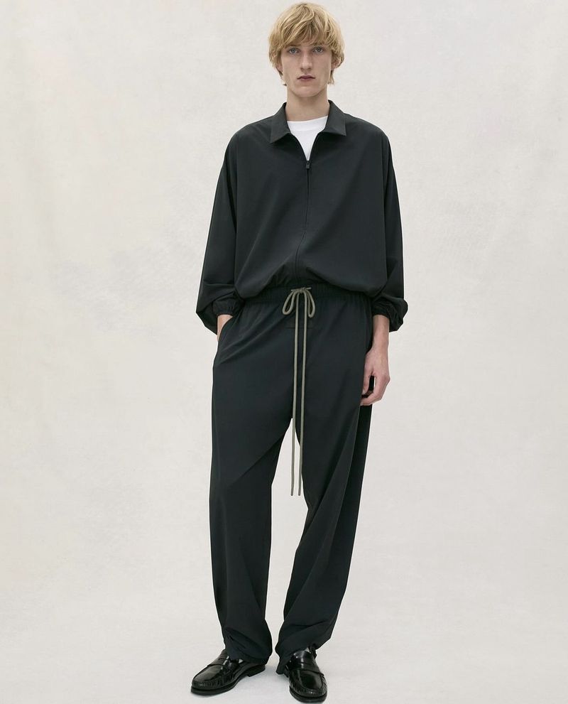 Full-length studio shot of a fair-skinned male model in a black matching top and bottom outfit with silver drawstrings and black dress shoes.