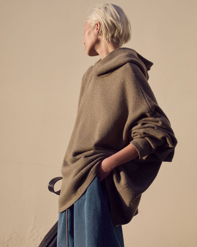 Woman in oversized brown hoodie and blue sweatpants stands outside in soft, natural light. Fashion shot. Outdoor setting.