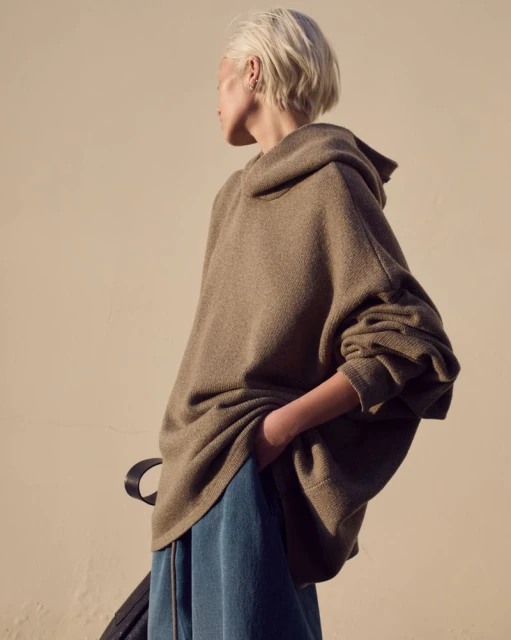 Woman in oversized brown hoodie and blue sweatpants stands outside in soft, natural light.  Fashion shot. Outdoor setting.