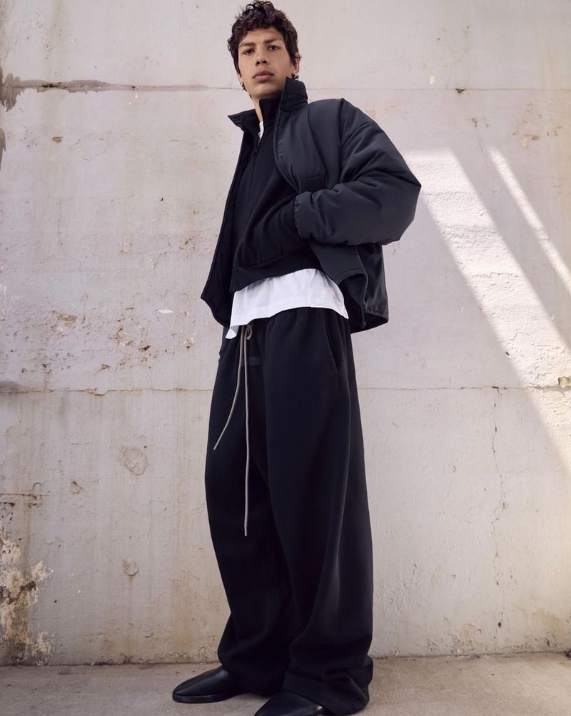 Man in a black puffer jacket, sweater, white undershirt, and black pants stands against an off-white textured wall in a casual pose.