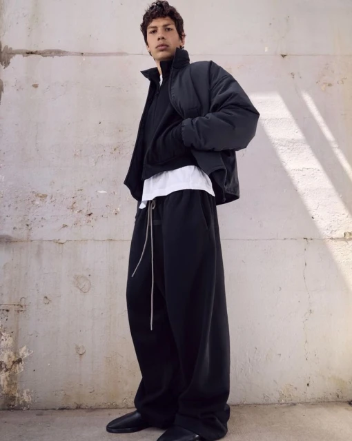 Man in a black puffer jacket, sweater, white undershirt, and black pants stands against an off-white textured wall in a casual pose.