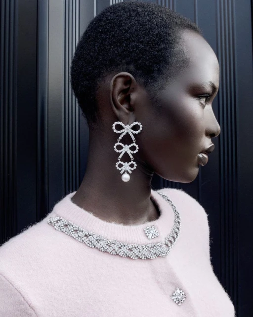 Profile shot of a Black female model wearing a pink cardigan, elegant diamond necklace and bow earrings against a dark background.