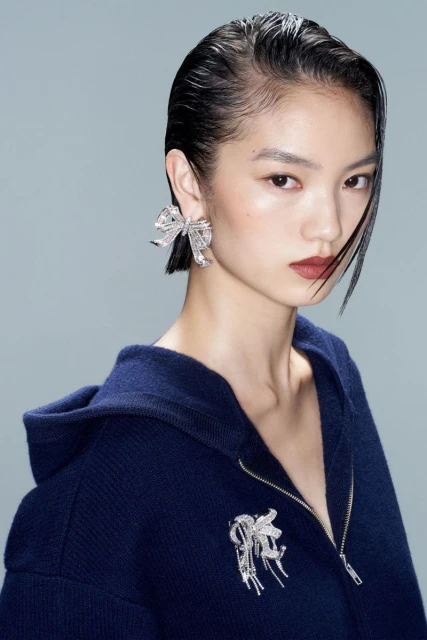 Portrait of an Asian woman with short hair wearing a navy blue hooded jacket, adorned with crystal brooch and bow earrings.