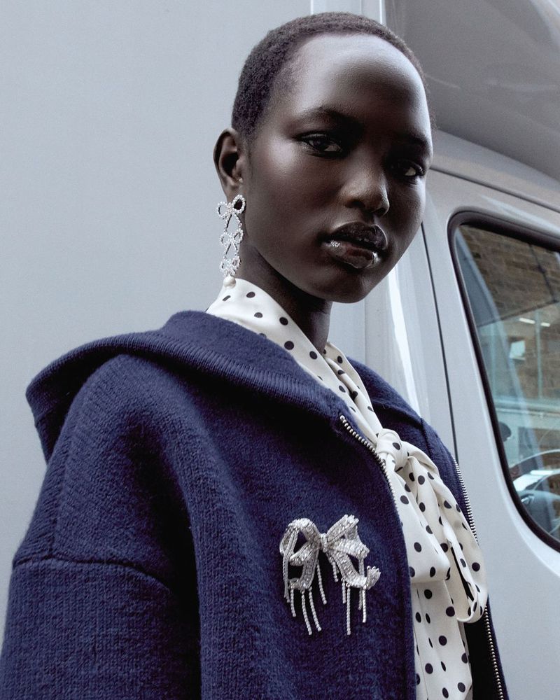 Portrait of a Black woman in a navy jacket with a bow brooch and bow earrings, wearing a white polka dot blouse, standing outdoors.