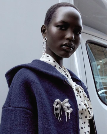 Portrait of a Black woman in a navy jacket with a bow brooch and bow earrings, wearing a white polka dot blouse, standing outdoors.