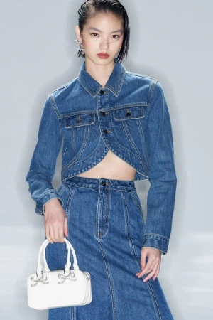 Model wearing a cropped blue denim jacket and matching skirt holding a small white handbag against a pale grey studio background.