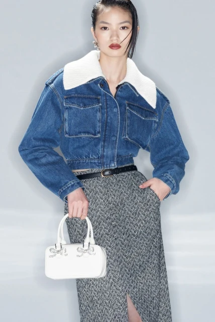 Stylish woman poses in a denim jacket, patterned skirt, and white handbag against a neutral backdrop, showcasing fashion and elegance.